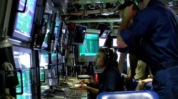 People in navy uniforms operate sonar and computer screens inside a submarine control room, surrounded by monitors and technical equipment in low lighting.