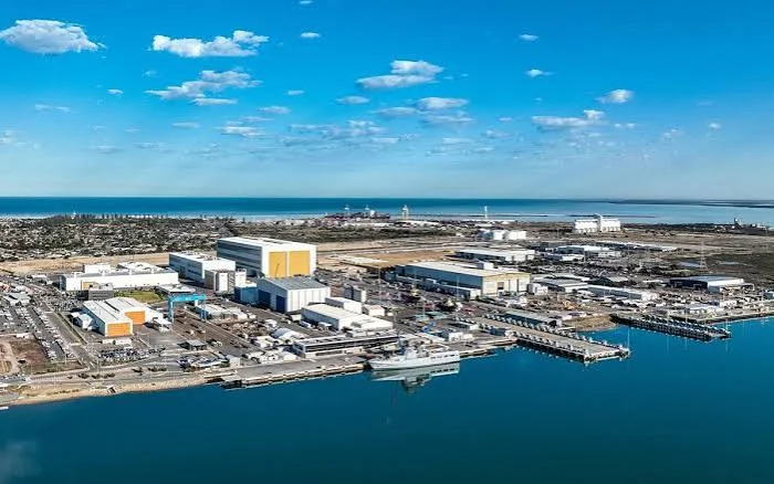 Aerial view of an industrial port facility by the water, with large warehouses, docks, ships, and a clear blue sky.