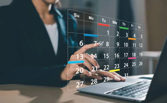 A person using a laptop interacts with a transparent digital calendar, selecting a date. The calendar displays weeks, with colorful lines highlighting specific days and events.