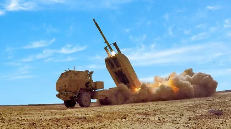 A military truck-mounted missile launcher fires a missile, producing clouds of dust and smoke in a desert landscape under a clear blue sky.