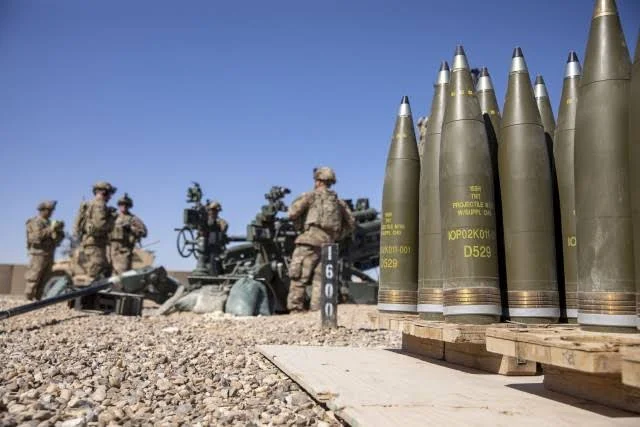 Several artillery shells are lined up on wooden pallets in the foreground, with a group of soldiers and a large artillery gun in the background under a clear blue sky.