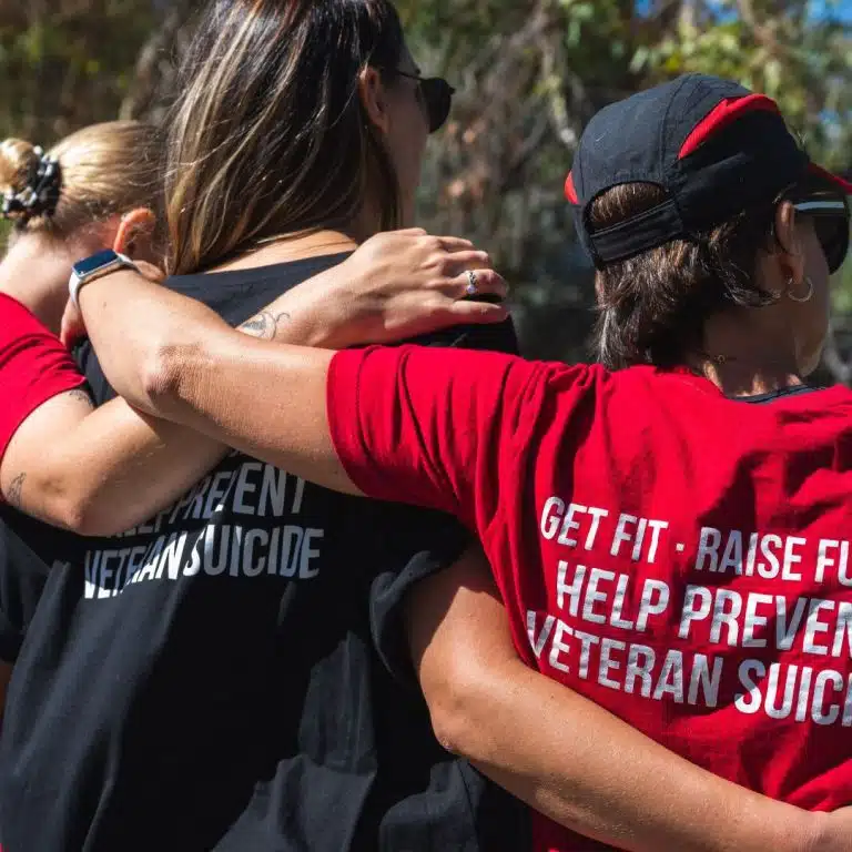 People stand outdoors with arms around each other. One person wears a red shirt that says “GET FIT. HELP PREVENT VETERAN SUICIDE.” The scene suggests unity and support at an awareness or fundraising event.
